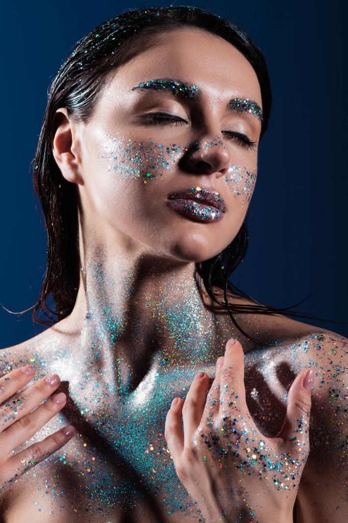 portrait of young woman with closed eyes and sparking glitter on body isolated on blue  - Photo, Image