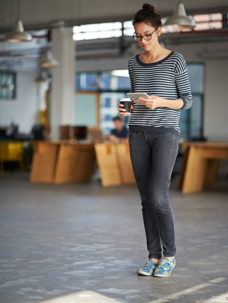 Техніка підтримки її зв'язку. Молода жінка тримає каву і дивиться на свій цифровий планшет у відкритому офісі
 - Фото, зображення