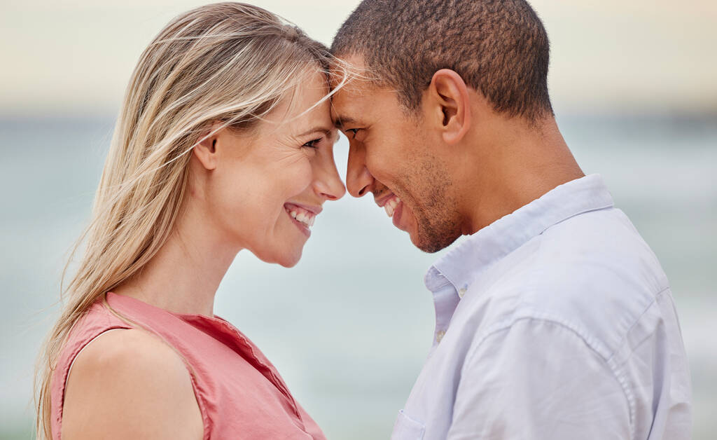 Portret van een stel op het strand met hun voorhoofd ontroerend. Multiraciale man en vrouw in relatie, gelukkig, glimlachend en verliefd op vakantie. Verbinding, liefde en kwaliteitstijd samen. - Foto, afbeelding