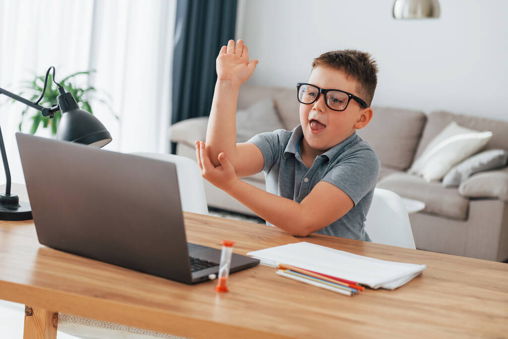 Σήκωσε το χέρι του. Μικρό αγόρι έχει online μαθήματα με τη χρήση φορητού υπολογιστή στο σπίτι. - Φωτογραφία, εικόνα