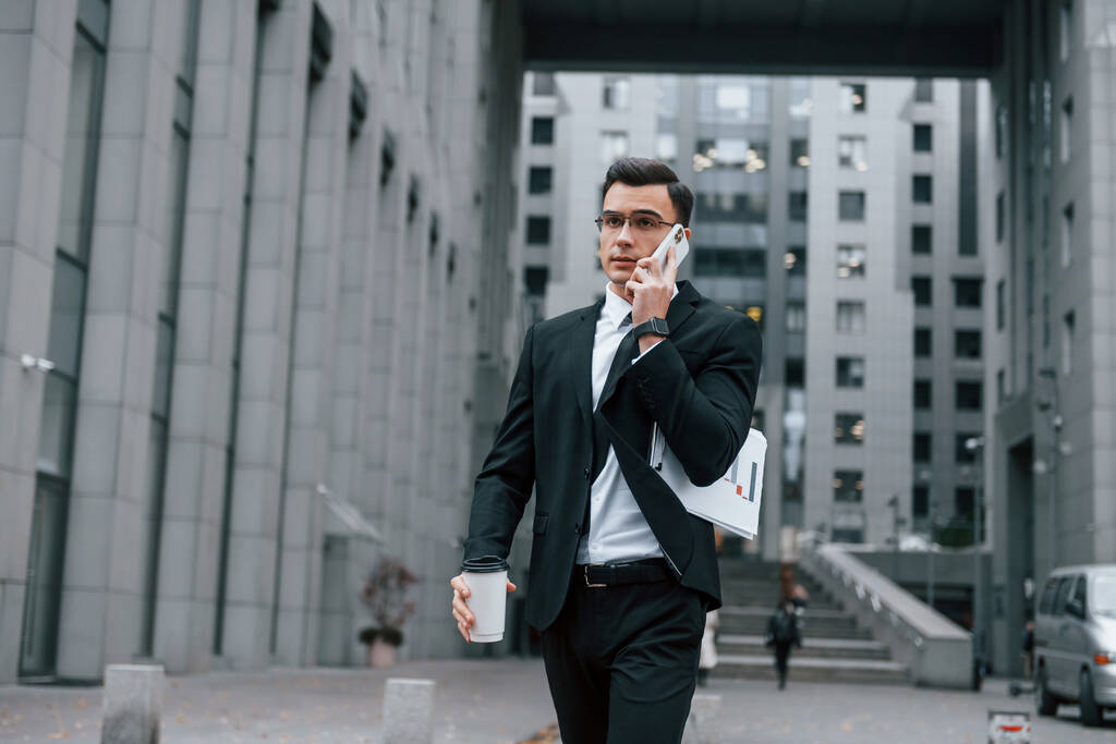 Walking and talking by phone. Businessman in black suit and tie is outdoors in the city. - Photo, Image
