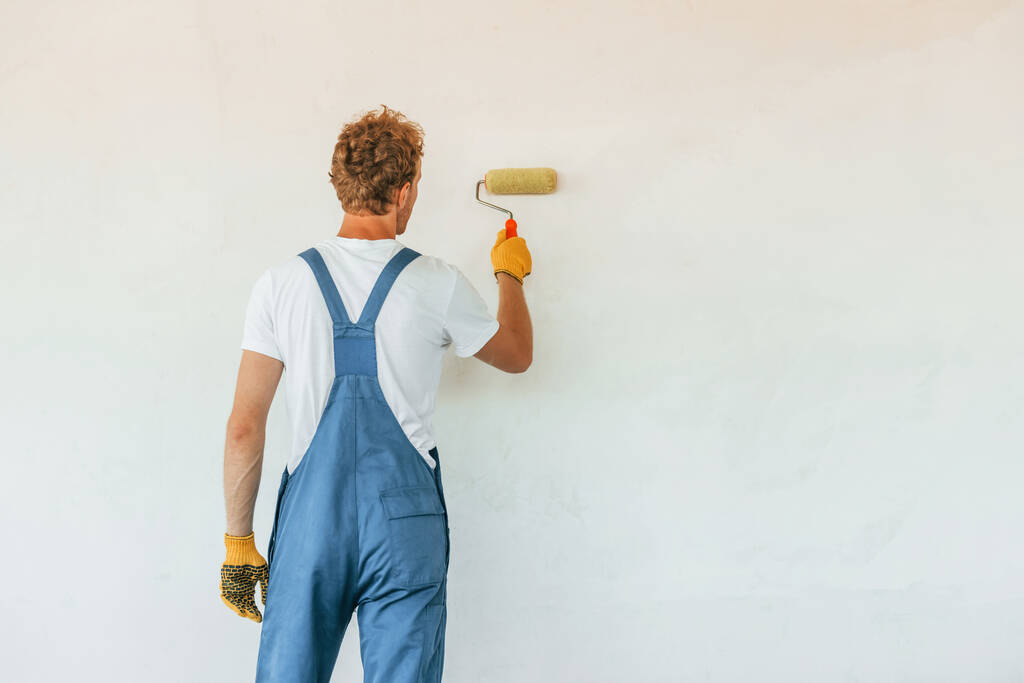 Покраска стен. Молодой человек, работающий в форме на стройке днем. - Фото, изображение