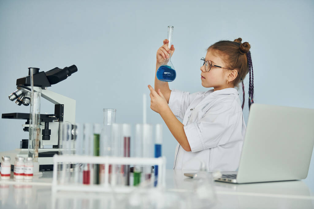 Palto giyen küçük bir kız laboratuarda ekipman kullanarak bilim adamını oynuyor.. - Fotoğraf, Görsel