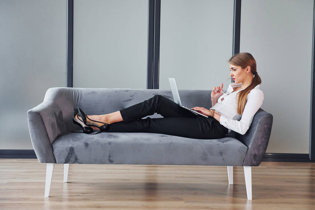 С ноутбуком. Молодая женщина в формальной одежде днем сидит на диване. - Фото, изображение