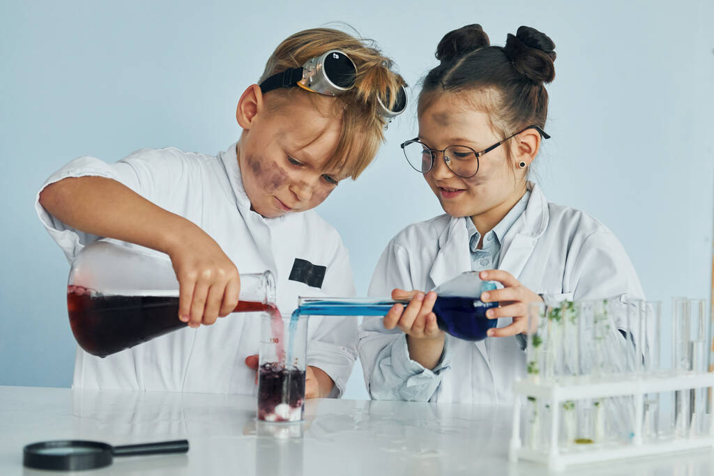 Маленькая девочка и мальчик в белых халатах играют ученых в лаборатории с помощью оборудования. - Фото, изображение