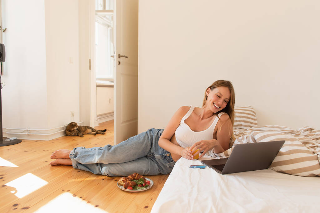 Junge Frau hält Orangensaft in der Nähe von Laptop und Frühstück im Schlafzimmer  - Foto, Bild