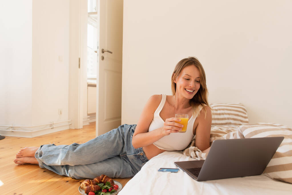Wesoła młoda kobieta trzyma sok pomarańczowy w pobliżu laptopa i śniadanie w sypialni  - Zdjęcie, obraz