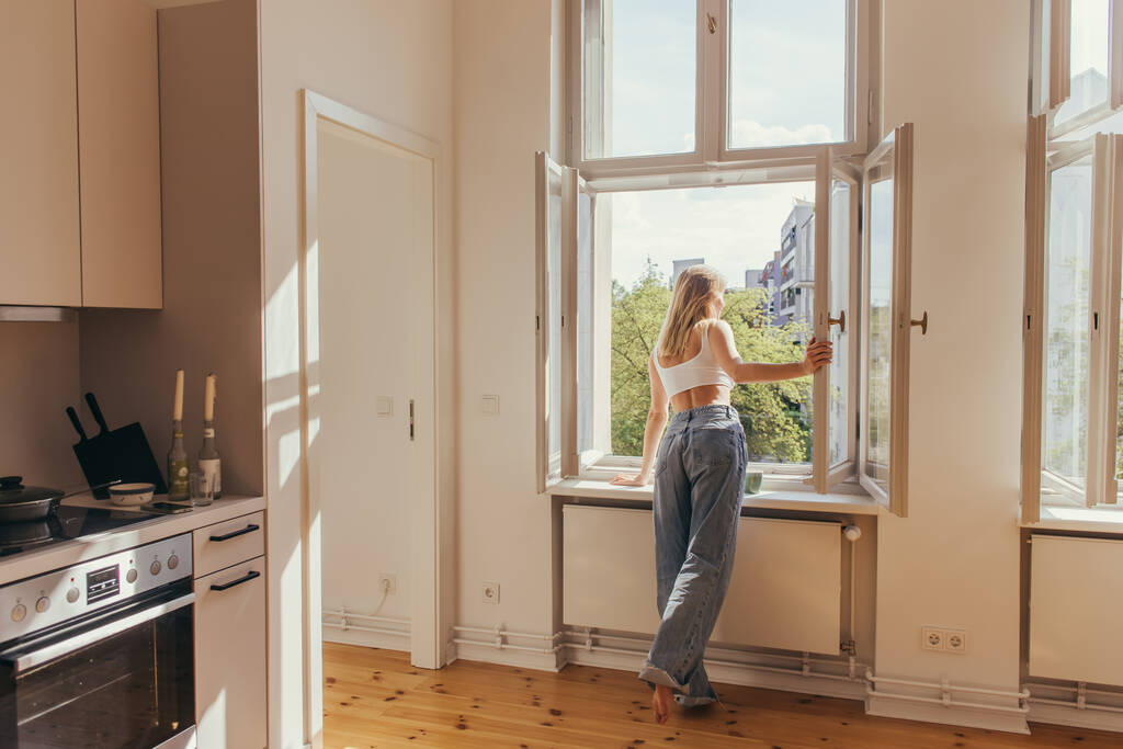 Блондинка смотрит на открытое окно возле чашки кофе на подоконнике на кухне  - Фото, изображение