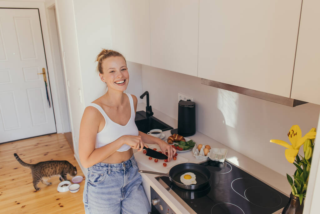 Mujer feliz mirando a la cámara mientras corta tomates cherry cerca del huevo en la sartén en la cocina  - Foto, Imagen