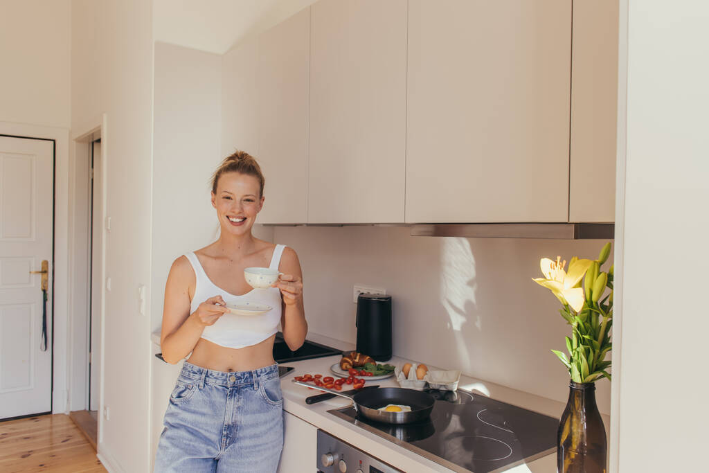 Mujer sonriente en la parte superior sosteniendo taza de café cerca del desayuno y flores en la cocina  - Foto, imagen