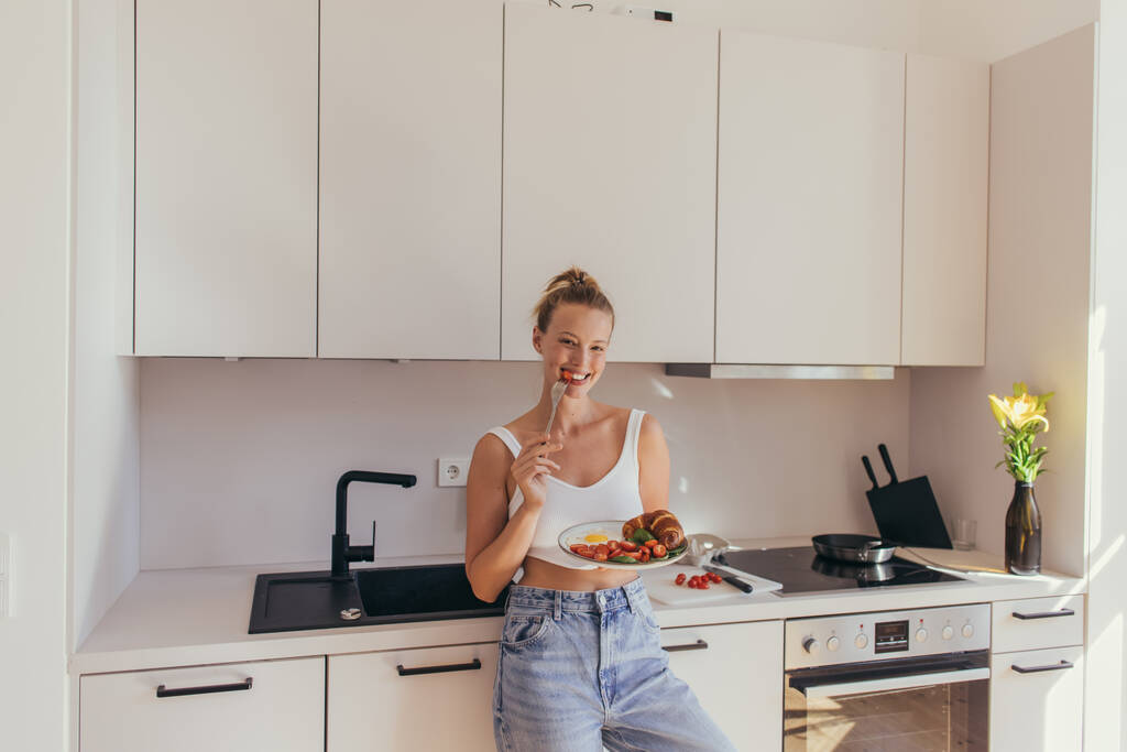Hymyilevä nainen syö kirsikkatomaattia pitäessään aamiaista keittiössä  - Valokuva, kuva