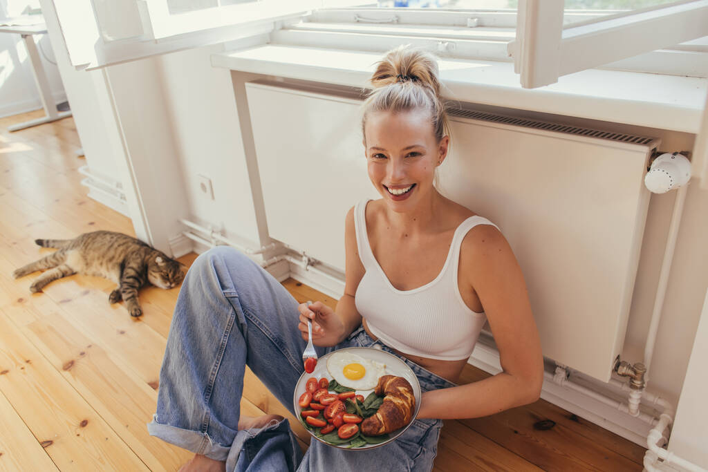 Mujer sonriente en la parte superior del plato de celebración con sabroso desayuno cerca borrosa gato pliegue escocés en casa  - Foto, Imagen
