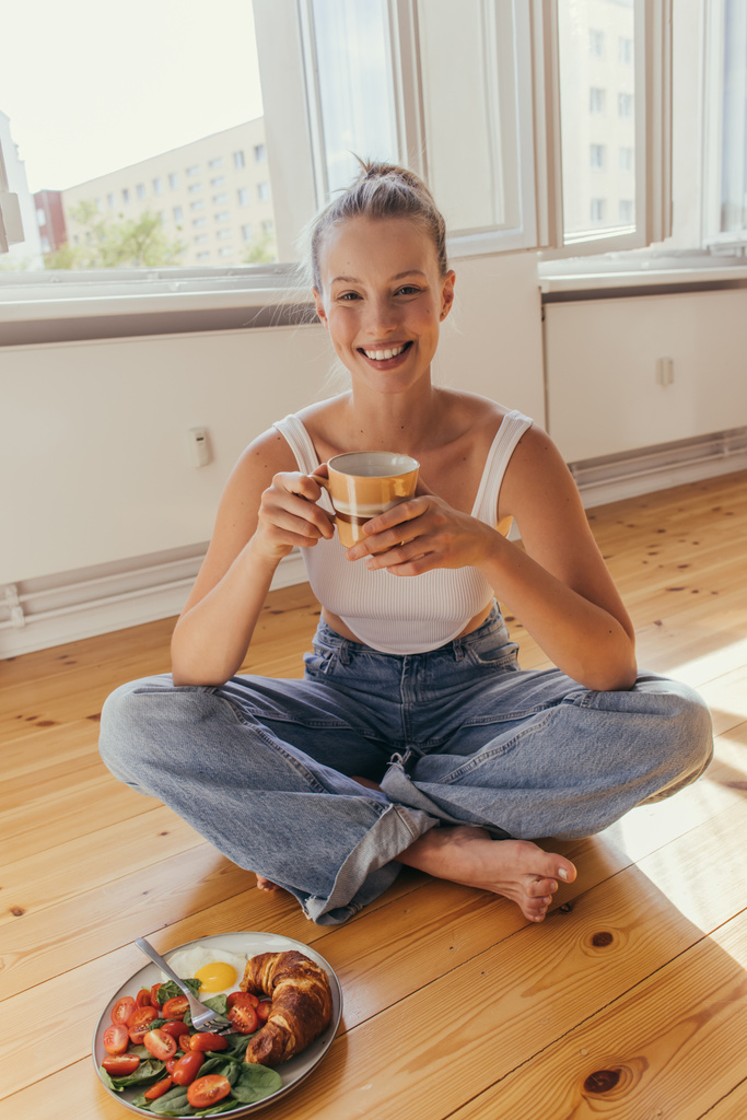 Donna bionda sorridente in jeans e tazza di tenuta superiore vicino a una gustosa colazione sul pavimento a casa  - Foto, immagini