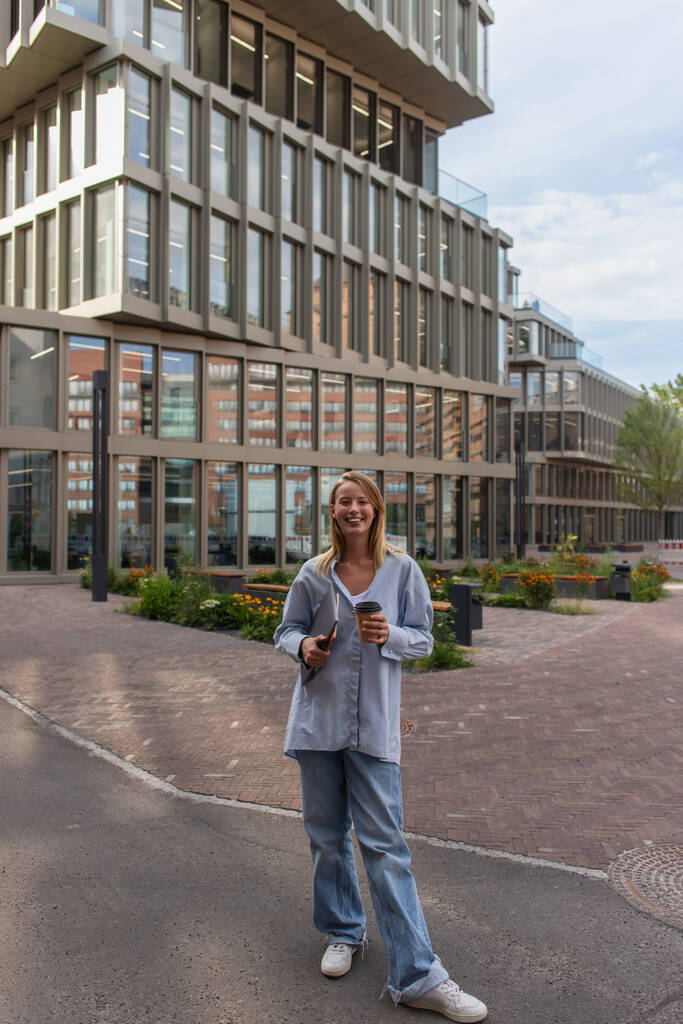 Usmívající se žena drží kávu jít a notebook na městské ulici v Berlíně  - Fotografie, Obrázek