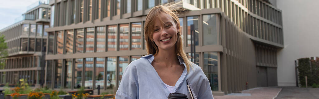Joven alegre sosteniendo portátil y café para ir cerca de edificio borroso en la calle en Berlín, pancarta  - Foto, imagen