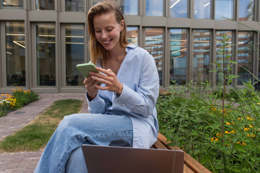 Lächelnder Freiberufler mit Smartphone in Laptopnähe auf Bank im Freien  - Foto, Bild