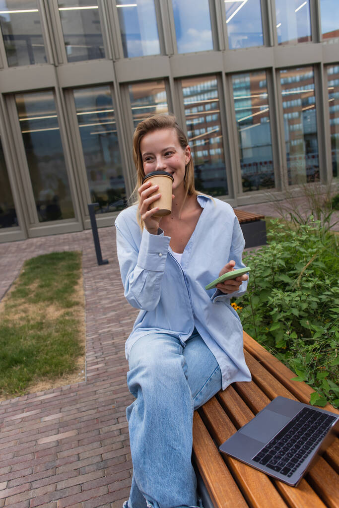 Young woman holding smartphone and drinking coffee to go near laptop on bench on street  - Photo, Image