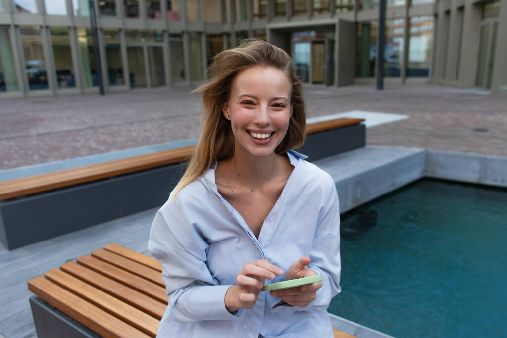 Cheerful blonde woman in shirt holding smartphone and looking at camera on bench on street  - Photo, Image