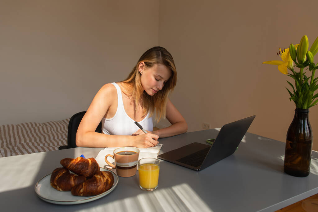 Libero professionista che scrive su taccuino vicino a colazione con bevande e gadget a casa  - Foto, immagini