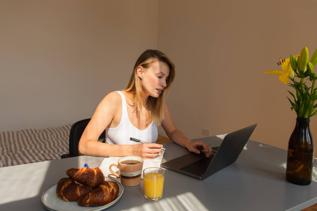 Giovane freelance con computer portatile e notebook vicino a colazione con bevande a casa  - Foto, immagini