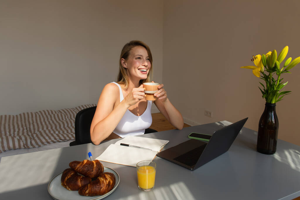 Счастливая блондинка, держащая чашку за завтраком и гаджеты дома  - Фото, изображение