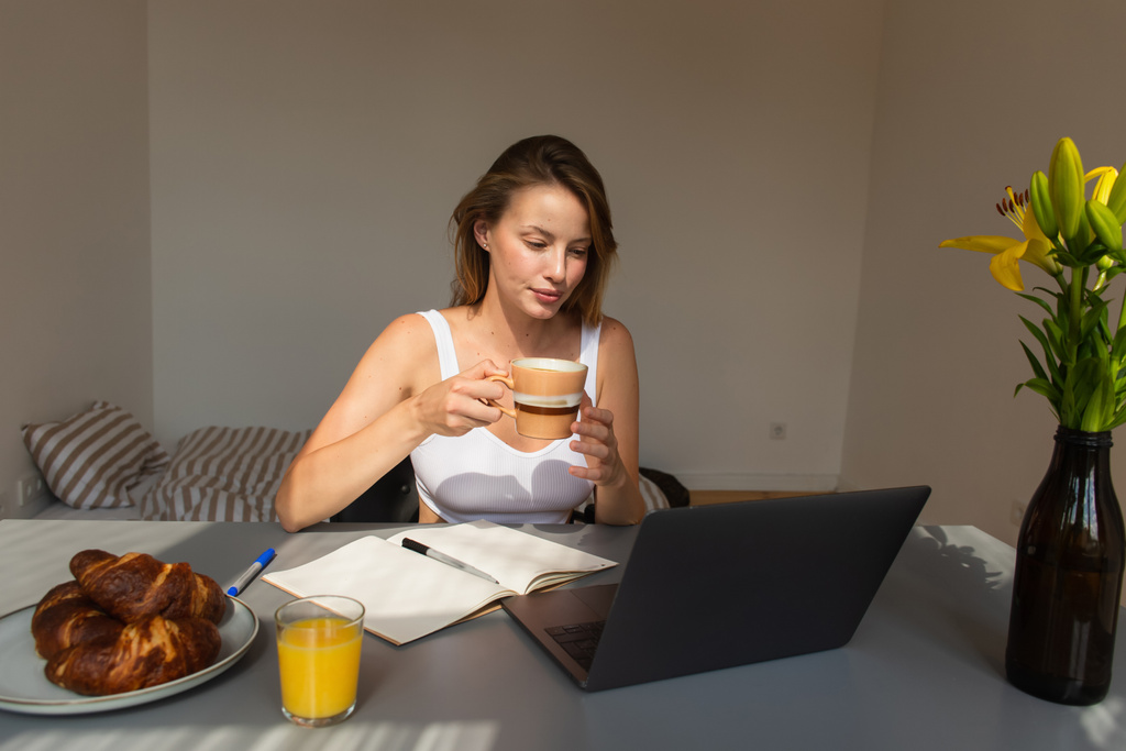 Blond vrouw met beker en kijken naar laptop in de buurt van bloemen en ontbijt thuis  - Foto, afbeelding