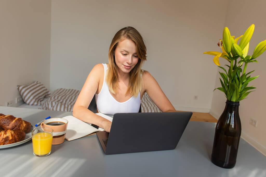 Mujer rubia usando el ordenador portátil cerca del desayuno y bebidas en casa  - Foto, Imagen