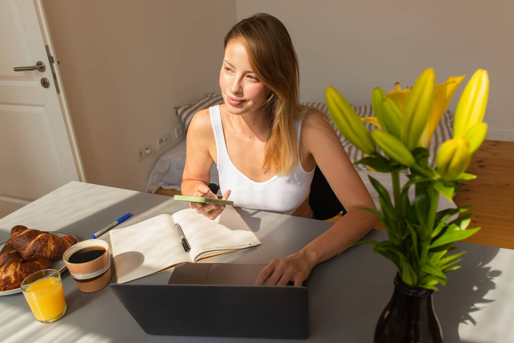 Blondynka freelancer za pomocą smartfona i laptopa w pobliżu śniadania w domu  - Zdjęcie, obraz