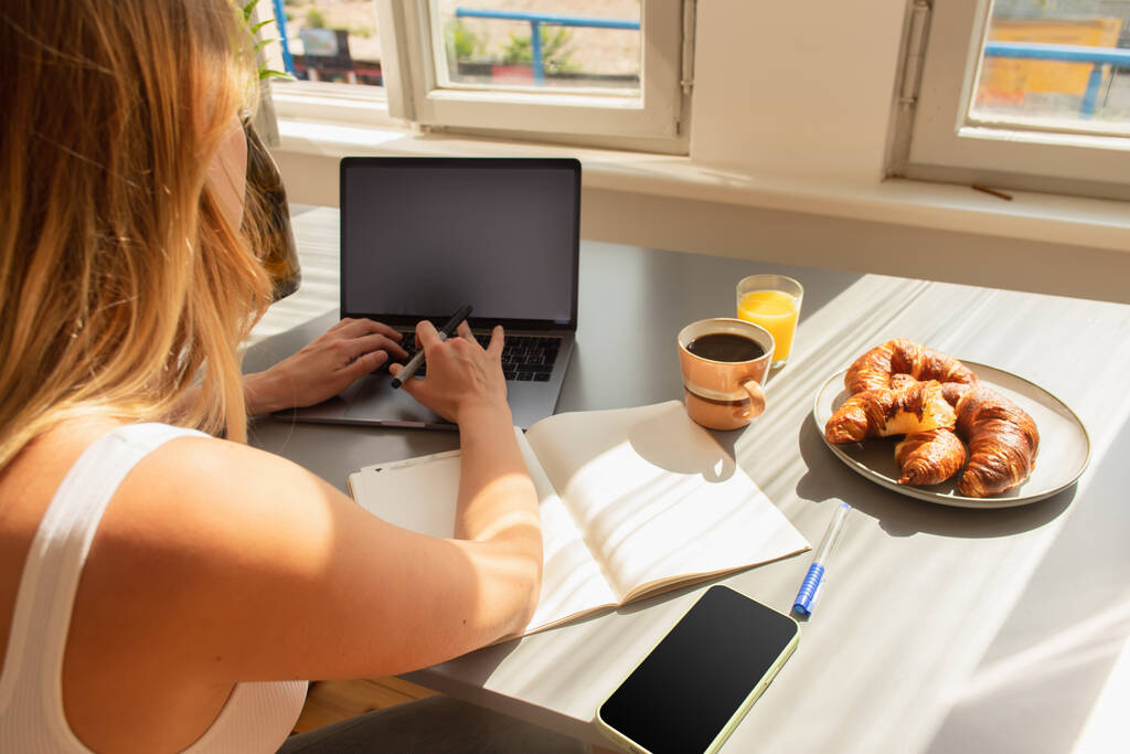 Jeune femme utilisant un ordinateur portable près d'un ordinateur portable et petit déjeuner sur la table à la maison  - Photo, image