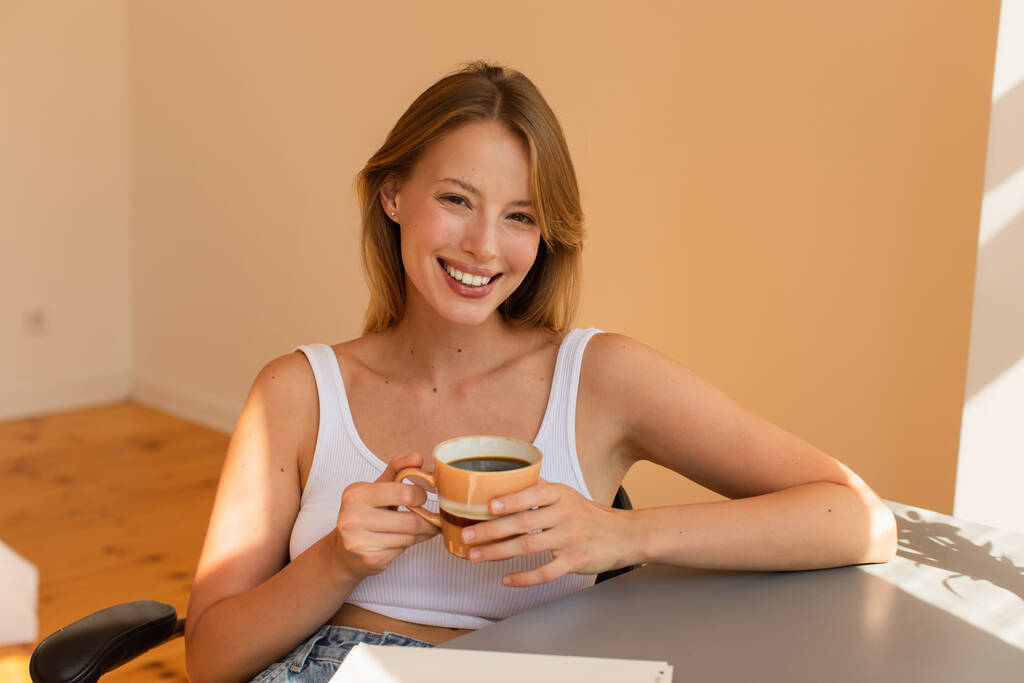 Mulher loira alegre segurando xícara de café perto notebook na mesa em casa  - Foto, Imagem