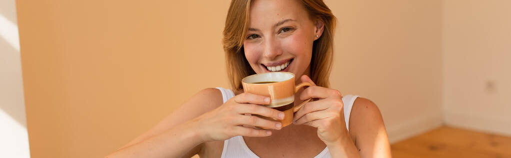 Mulher loira positiva segurando xícara de café e olhando para a câmera em casa, banner  - Foto, Imagem