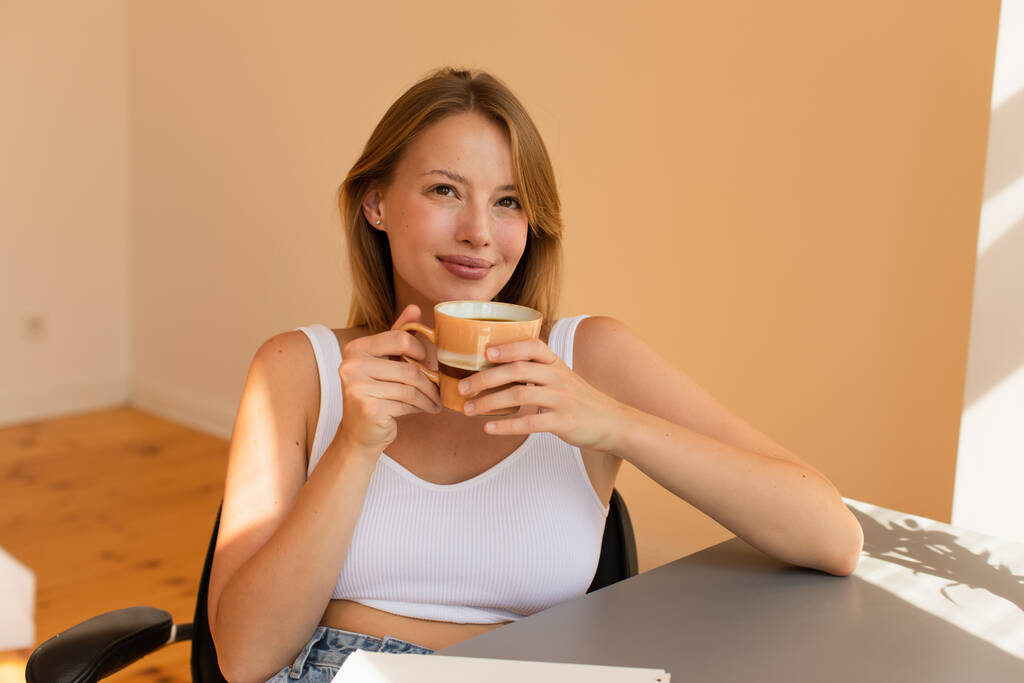 Мечтательная блондинка в верхней держать чашку кофе дома  - Фото, изображение