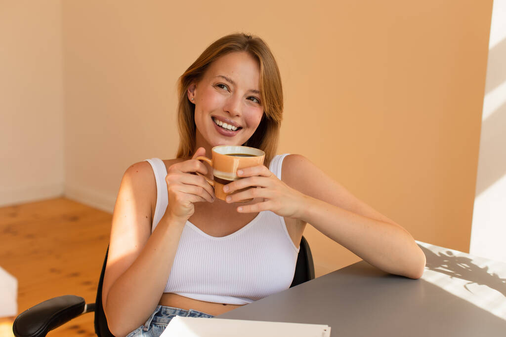 Veselá mladá žena v horní drží šálek kávy u stolu doma  - Fotografie, Obrázek