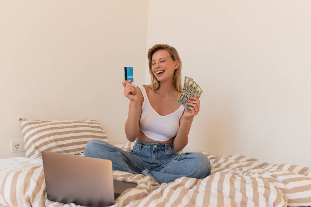 Blonde Frau lacht, während sie Bargeld und Kreditkarte in der Nähe von Laptop auf dem Bett hält  - Foto, Bild