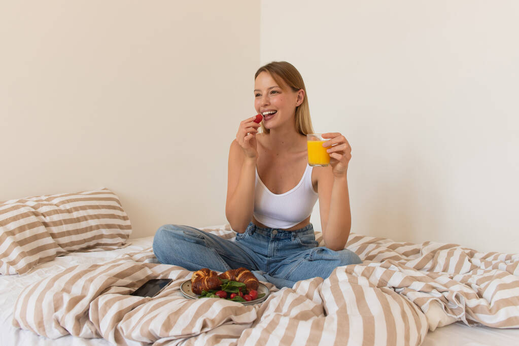 Mujer sonriente sosteniendo tomate cherry y jugo de naranja cerca de croissants y smartphone en la cama  - Foto, Imagen