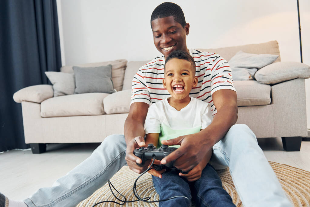 Con joysticks en las manos. Padre afroamericano con su hijo pequeño en casa. - Foto, Imagen