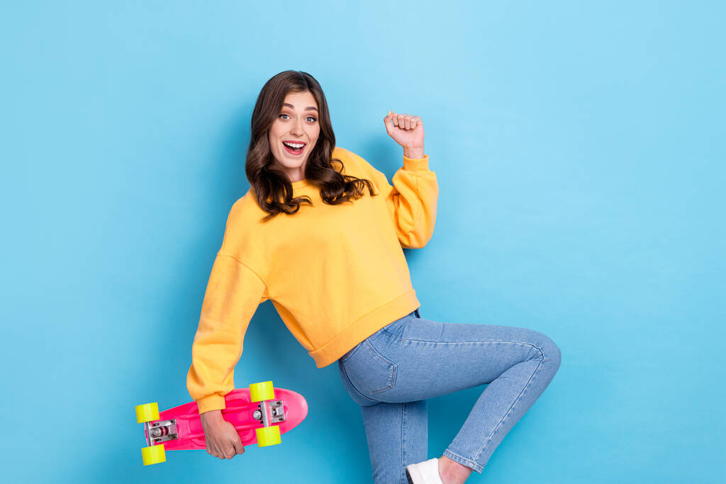 Фотопортрет милої молодої жінки, що тримає скейтборд, піднімає кулак, святкує продаж одягу модного жовтого кольору, ізольованого на синьому фоні
. - Фото, зображення