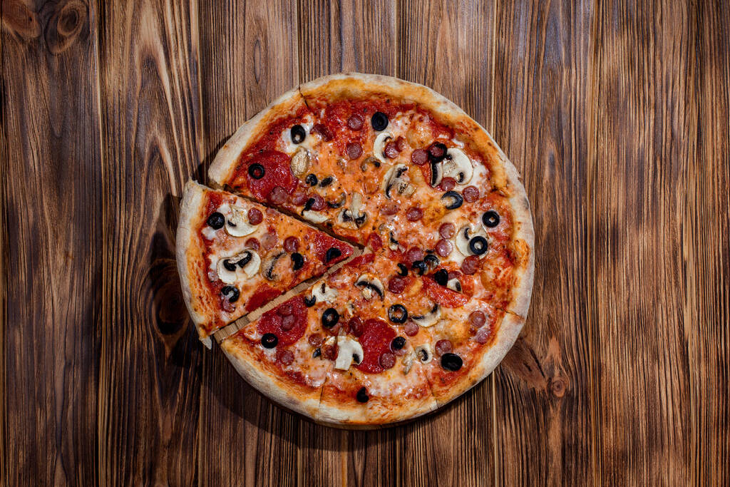 Tomato-based pizza with mozzarella cheese, hunting sausages, salami, olives, mushroom sauce. On a wooden background. - Zdjęcie, obraz
