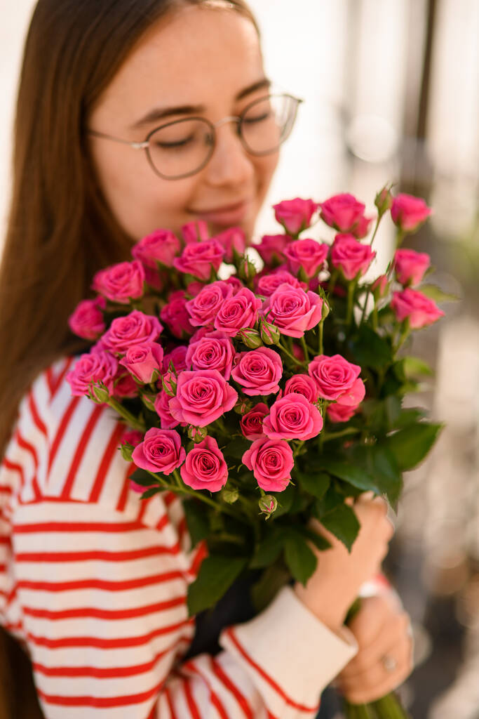 Vista da vicino di giovane donna sorridente teneramente in mano e abbracciando mazzo di rose rosa brillante. Ritratto di ragazza con fiori - Foto, immagini