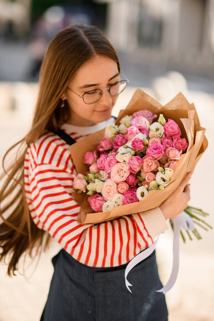 Selective focus on bouquet of pink rose flowers in wrapping paper which tenderly holding young woman and look at it - Photo, Image