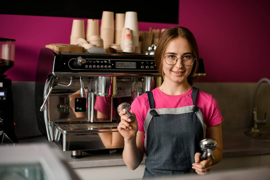 portrait d'une jolie jeune femme barista dans des lunettes et tablier dans un café. Machine à café moderne professionnelle en arrière-plan - Photo, image