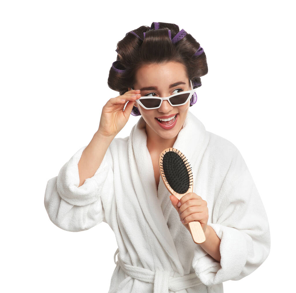 Fröhliche junge Frau im Bademantel mit Lockenwicklern und Sonnenbrille, die zur Haarbürste auf weißem Hintergrund singt - Foto, Bild
