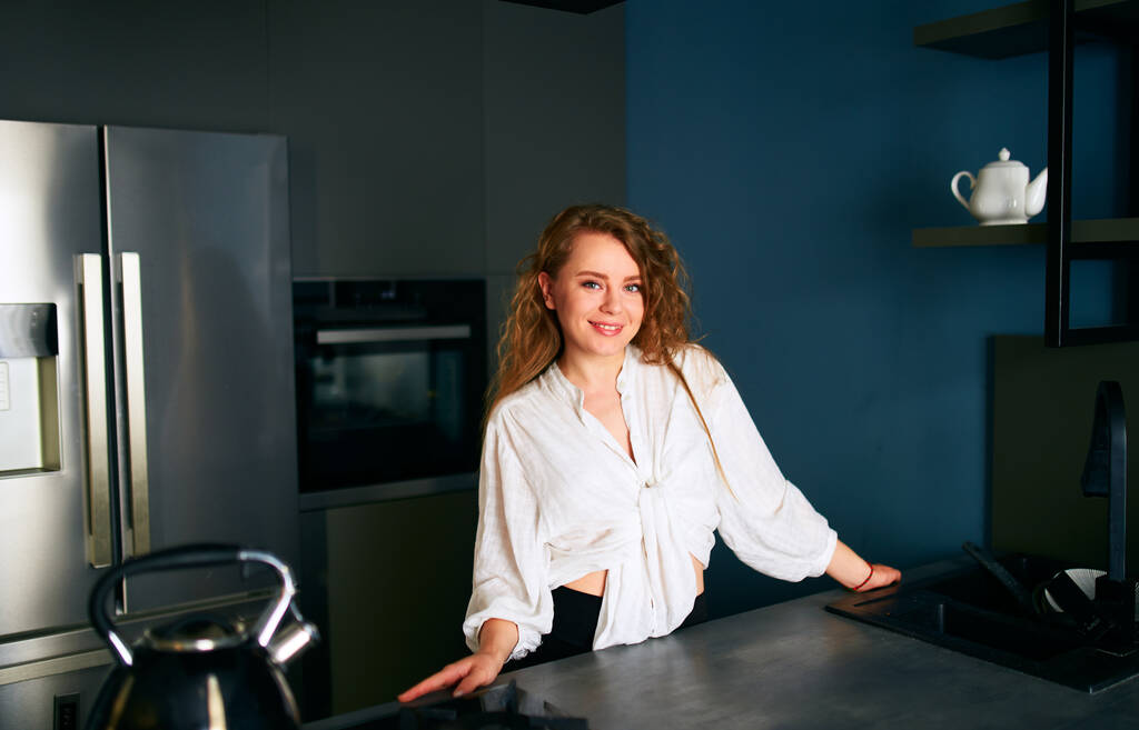 Portrait of a blonde caucasian woman standing and smiling in her kitchen leaning on the table. Young cheerful adult female standing at home in modern interior with hands on cooking surface - Fotografie, Obrázek