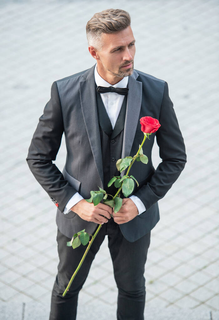 елегантна людина в тукседо. чоловік у чорному костюмі на відкритому повітрі. красивий смокінг чоловік з червоною трояндою
. - Фото, зображення
