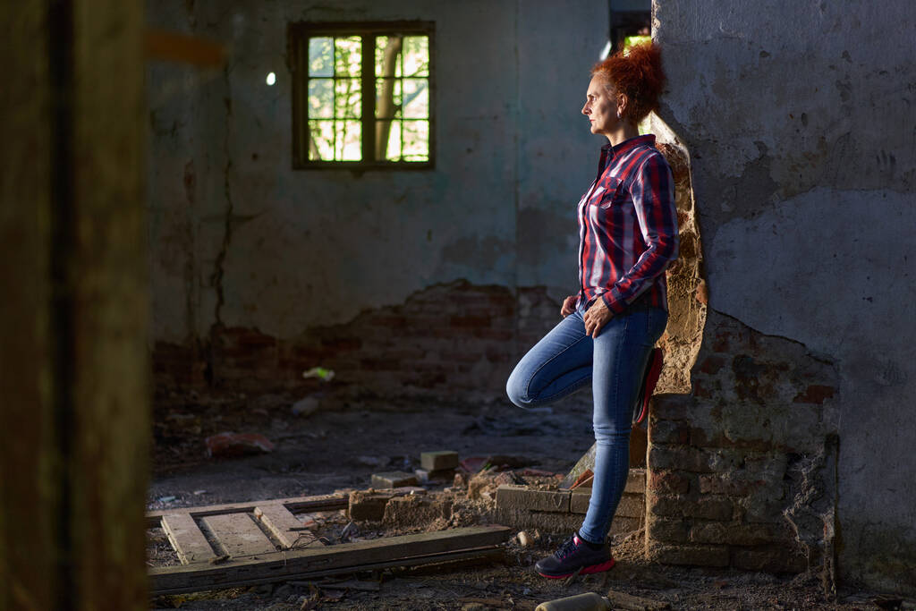 Femme rousse aux cheveux bouclés en chemise à carreaux et jeans bleus dans une grange et une maison en ruine - Photo, image