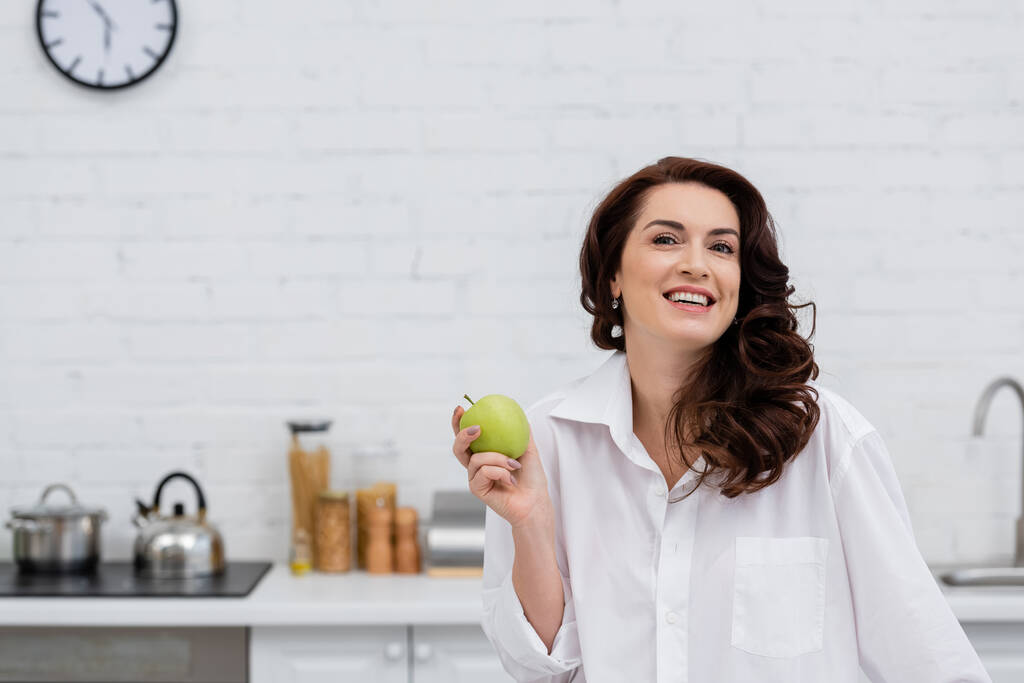 Χαμογελαστή γυναίκα με πουκάμισο κρατώντας φρέσκο μήλο στην κουζίνα - Φωτογραφία, εικόνα