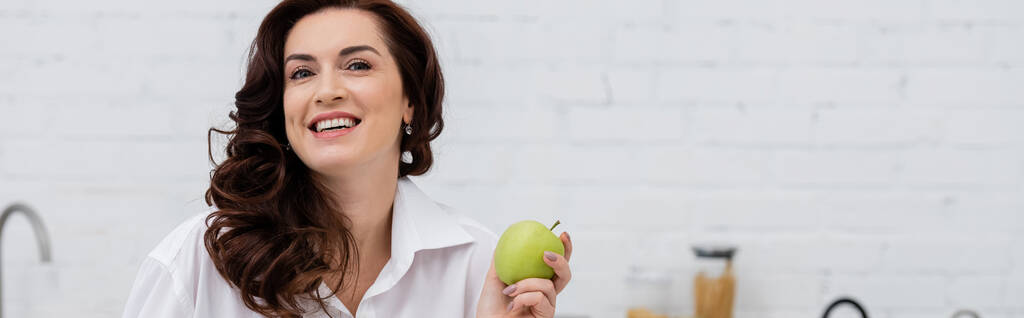 Позитивная брюнетка в рубашке держит спелое яблоко на кухне, баннер  - Фото, изображение