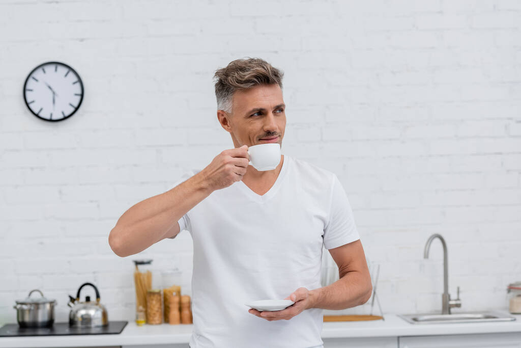 Homme en t-shirt tenant une tasse de café dans la cuisine le matin  - Photo, image