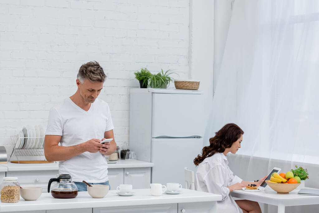 Couple utilisant des appareils près du petit déjeuner et du café dans la cuisine  - Photo, image