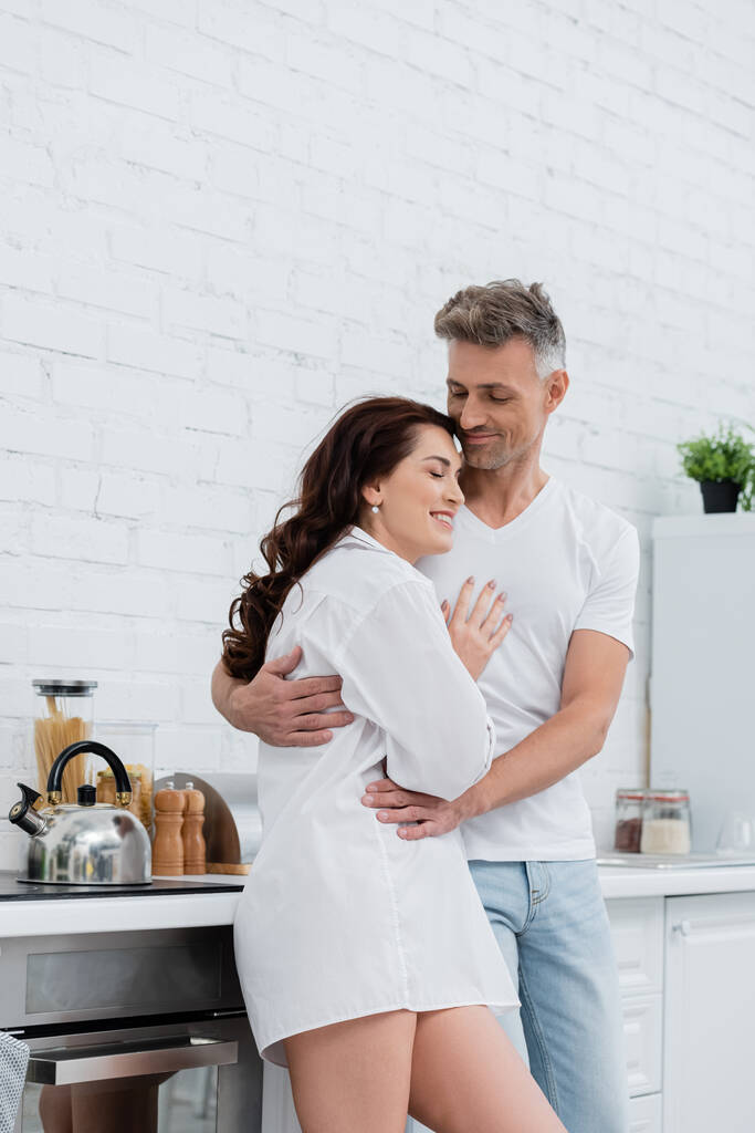 Χαμογελαστή άνδρας αγκαλιάζει μελαχρινή γυναίκα σε πουκάμισο στην κουζίνα  - Φωτογραφία, εικόνα